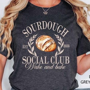 May include: Dark grey heather t-shirt with the text "SOURDOUGH SOCIAL CLUB Wake and bake" in a circular design with a bread loaf illustration. The shirt also has the text "EST 2026".