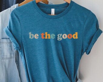 May include: A teal short-sleeved crew neck t-shirt with the text "be the good" printed in a multi-colored font featuring shades of light blue, orange, and yellow. The shirt is displayed on a wooden hanger alongside a light blue denim shirt.