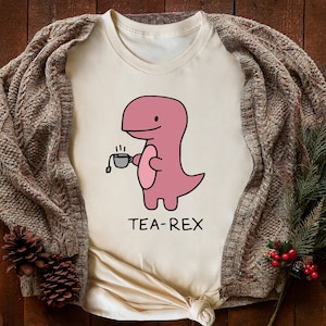 May include: A white t-shirt with a pink cartoon dinosaur holding a cup of tea. The text "TEA-REX" is printed below the dinosaur.