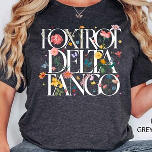 May include: Dark heather grey t-shirt with a floral design and the text "Fox Trot Delta Tango" in white.