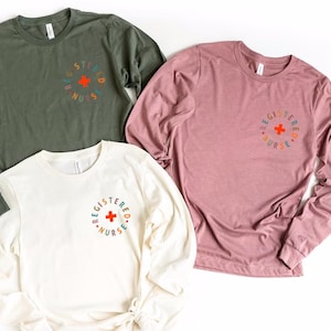 May include: Three long-sleeved shirts with the text "Registered Nurse" in a circle around a red cross. The shirts are in olive green, light pink, and cream.