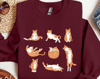 May include: A maroon crewneck sweatshirt featuring a graphic print of eight orange and white tabby cats in various poses, including sitting, stretching, rolling, and standing. The cats are illustrated in a simple, clean style across the center.