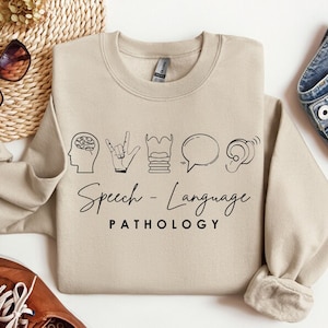 May include: A beige sweatshirt with a black graphic of a brain, a hand signing, a stack of books, a speech bubble, and an ear. The text "Speech - Language PATHOLOGY" is printed below the graphic.