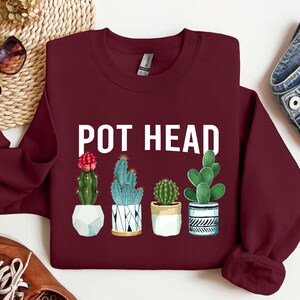 May include: Burgundy sweatshirt with the text "POT HEAD" in white lettering. The sweatshirt features a graphic of five potted cacti with different colors and patterns.