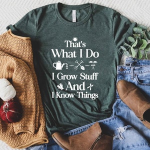 May include: A dark green t-shirt with white text that reads "That's What I Do I Grow Stuff And I Know Things". The text is accompanied by a sun, watering can, shovel, and plant icons. The t-shirt is laid out on a white surface with a brown sweater, blue jeans, and brown boots.