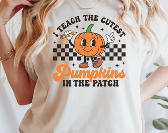 Puede incluir: Una camiseta de color crema con un gráfico de un personaje de calabaza de dibujos animados sosteniendo un lápiz. El texto dice I TEACH THE CUTEST PUMPKINS IN THE PATCH. El diseño incluye un patrón de cuadros blanco y negro y estrellas.