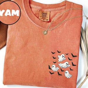 May include: Orange Comfort Colors t-shirt with a crew neck and short sleeves. The shirt features a graphic of three white ghosts and black bats. A gold necklace with a pendant is draped across the shirt. The word "YAM" is in a circle in the upper left corner.