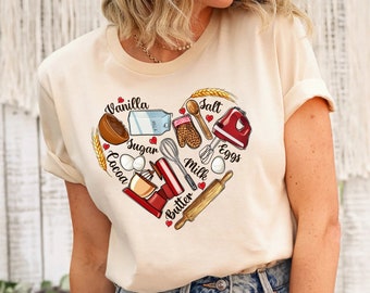 Könnte beinhalten: Ein beiges T-Shirt mit einem herzförmigen Design, das verschiedene Backzutaten und -werkzeuge zeigt. Das Design enthält die Wörter "Vanilla", "Salt", "Sugar", "Cocoa", "Eggs", "Milk" und "Butter".
