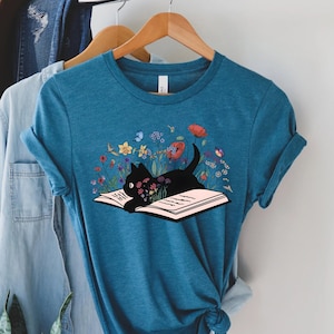 May include: A teal blue t-shirt with a black cat reading a book surrounded by colorful flowers. The cat is lying on the open book with its head resting on the pages.