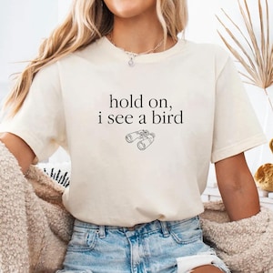 May include: Cream-colored t-shirt with the text "hold on, i see a bird" and a line drawing of binoculars. The shirt is paired with denim shorts. A cozy, cream-colored sweater is draped over the arms.