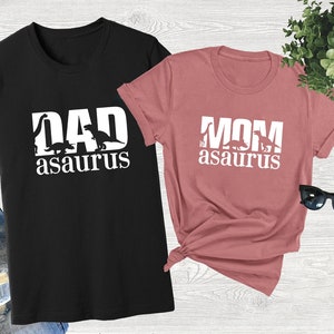 May include: A black t-shirt with the text "DAD asaurus" in white, featuring a silhouette of a dinosaur. A pink t-shirt with the text "MOM asaurus" in white, featuring a silhouette of a dinosaur.