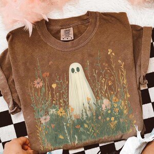 May include: Brown Comfort Colors t-shirt featuring a whimsical ghost design in a field of wildflowers. The ghost has a simple, friendly face. The shirt is a relaxed fit, perfect for casual wear. The shirt has the Comfort Colors brand tag.