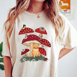 May include: A cream-colored t-shirt with a graphic of a cat sitting under a cluster of red and white mushrooms. The mushrooms have white spots and are surrounded by green grass and white flowers.