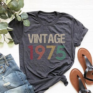May include: A dark gray t-shirt with the text "Vintage 1975" in colorful retro-style font.