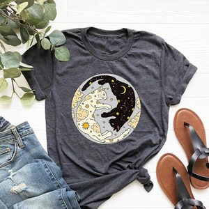 May include: A dark gray t-shirt with a graphic of two cats in a yin yang symbol. The cats are black and white and are surrounded by stars and clouds. The background of the graphic is a light gray.
