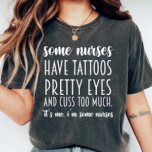 May include: A dark gray t-shirt with white text that reads "some nurses have tattoos pretty eyes and cuss too much. it's me. i'm some nurses". The shirt is worn by a person with long brown hair and a gold necklace.