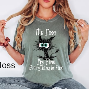 May include: Moss green t-shirt with a distressed graphic of a black cat with wide eyes and the text "It's Fine, I'm Fine, Everything Is Fine" in white. The shirt is a relaxed fit.