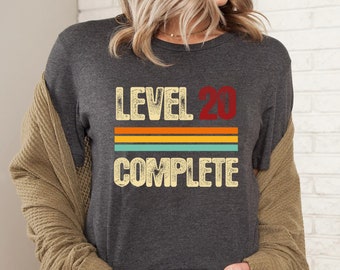 May include: A dark charcoal gray short-sleeve t-shirt featuring the text LEVEL 20 COMPLETE in a distressed, vintage-style font. The design includes horizontal stripes in orange, yellow, and teal, positioned between the words LEVEL 20 and COMPLETE.