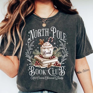 K&ouml;nnte beinhalten: Ein dunkelgraues T-Shirt mit einem wei&szlig;en Grafikdesign, das eine Tasse hei&szlig;er Schokolade mit Schlagsahne und einem Lebkuchenmann zeigt, umgeben von B&uuml;chern und Stechpalmen. Der Text lautet "North Pole Book Club Hot Cocoa Served Daily".