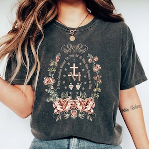 Op de afbeelding: Een donkergrijs T-shirt met een bloemenmotief en de tekst "O Maria, ontvangen zonder zonde, bid voor ons die onze toevlucht tot u nemen." Het ontwerp bevat een kruis, een hart en een kroon.
