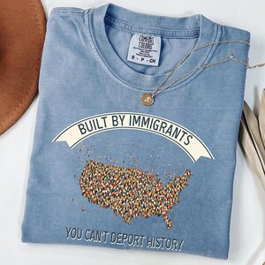 May include: A blue Comfort Colors t-shirt with the text "BUILT BY IMMIGRANTS" above a map of the United States filled with tiny figures. The phrase "YOU CAN'T DEPORT HISTORY" is printed below the map. A gold necklace is draped across the shirt.