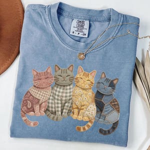 May include: A light blue Comfort Colors t-shirt featuring four smiling cat illustrations. Each cat has a unique patchwork design in shades of pink, gray, yellow, and blue. A gold necklace with a pendant rests on the shirt.
