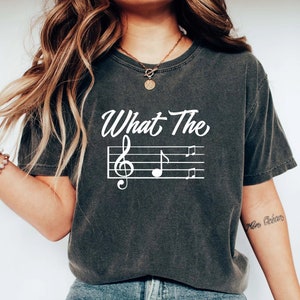 May include: A dark gray t-shirt with the text "What The" and a musical note staff with treble clef and notes printed in white.