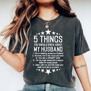 May include: A dark gray t-shirt with white text that reads "5 THINGS YOU SHOULD KNOW ABOUT MY HUSBAND" and a list of five things about a husband. The text is in a casual, humorous font.