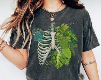 May include: A charcoal grey short-sleeve t-shirt featuring a graphic print of a human rib cage intertwined with various green tropical leaves, including monstera and palm fronds. The shirt has a crew neckline and a relaxed, vintage-washed finish.