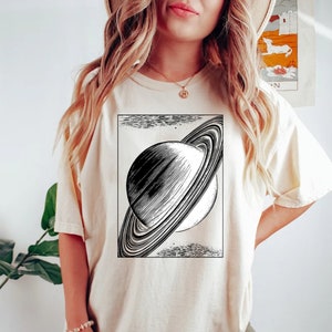 May include: A cream colored t-shirt with a black and white illustration of Saturn with its rings.