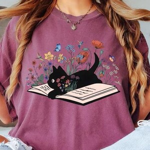 May include: A purple t-shirt with a black cat reading a book surrounded by flowers. The cat is sitting on an open book with the text "Berry" in the top right corner of the image.