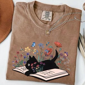 May include: A brown t-shirt with a black cat reading a book surrounded by flowers. The cat is sitting on an open book with a pink interior. The flowers are colorful and include blue butterflies. The text on the shirt reads "Comfort Colors S-P-CH".