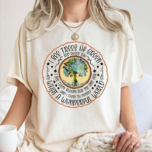 May include: A white t-shirt with a circular graphic featuring a tree with green leaves and a brown trunk. The text around the tree reads "I see trees of green, red roses too, I see them bloom for me and you, and I think to myself, what a wonderful world."