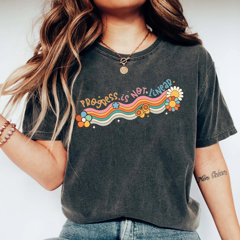 K&ouml;nnte beinhalten: Ein dunkelgraues T-Shirt mit einem Regenbogen-Motiv und dem Text "Progress is not linear". Das Motiv zeigt Blumen, Schmetterlinge und Sterne.