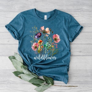 May include: A teal blue t-shirt featuring a watercolour floral design and the word "wildflower" printed in white.
