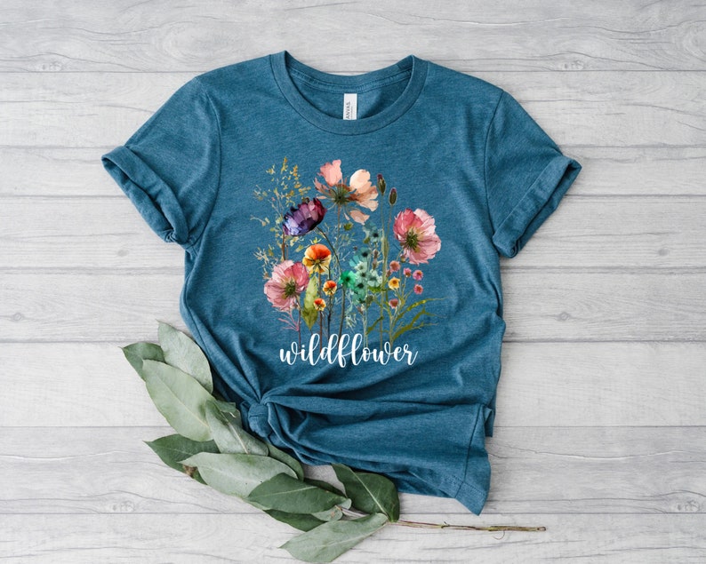 May include: A teal blue t-shirt featuring a watercolour floral design and the word "wildflower" printed in white.