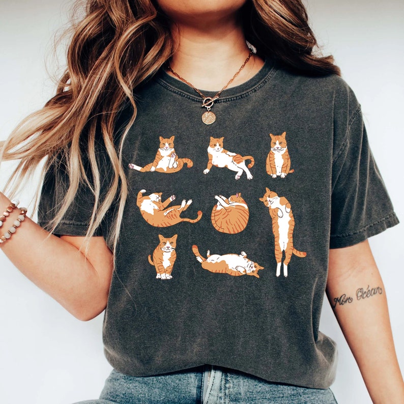 K&ouml;nnte beinhalten: Ein dunkelgraues T-Shirt mit einem Grafikdruck von orangefarbenen und wei&szlig;en Katzen in verschiedenen Yoga-Posen. Der Text "Mon Oc&eacute;ans" ist auf dem rechten Arm sichtbar.