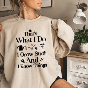 May include: A beige sweatshirt with black text that reads "That's What I Do I Grow Stuff And I Know Things". The text is accompanied by a sun, watering can, shovel, and plant icons.