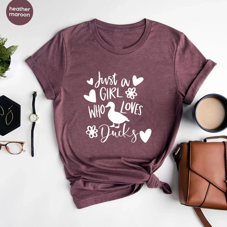 May include: Heather maroon t-shirt with the text "Just a GIRL who loves DUCKS" in white. The design includes hearts, flowers, and a duck illustration. The shirt is a short-sleeved crew neck.