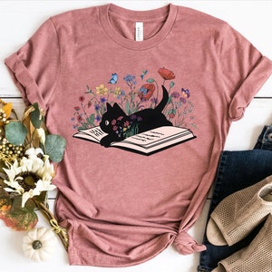 May include: A pink t-shirt with a black cat reading a book surrounded by colorful flowers. The cat is lying on the open book with its head resting on the pages. The flowers are a variety of colors, including red, pink, yellow, and blue.