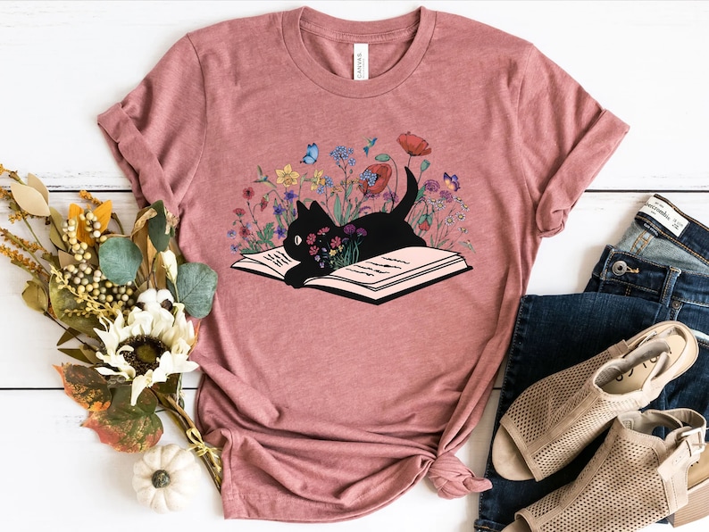 May include: A pink t-shirt with a black cat reading a book surrounded by colorful flowers. The cat is lying on the open book with its head resting on the pages. The flowers are a variety of colors, including red, pink, yellow, and blue.