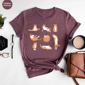K&ouml;nnte beinhalten: Ein T-Shirt in Heide-Maroon mit einem Aufdruck von orangefarbenen Tabby-Katzen in verschiedenen Yoga-Posen. Das Shirt hat einen Rundhalsausschnitt und kurze &Auml;rmel.