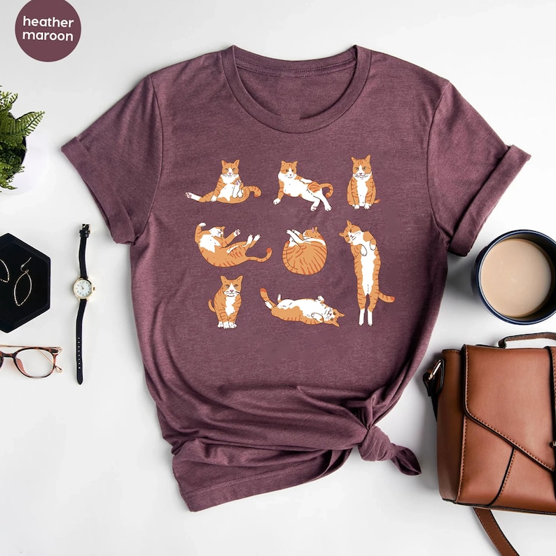 K&ouml;nnte beinhalten: Ein T-Shirt in Heide-Maroon mit einem Aufdruck von orangefarbenen Tabby-Katzen in verschiedenen Yoga-Posen. Das Shirt hat einen Rundhalsausschnitt und kurze &Auml;rmel.