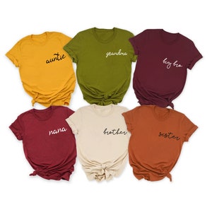 Peut inclure: Six t-shirts de diff&eacute;rentes couleurs avec des titres de membres de la famille imprim&eacute;s dessus en &eacute;criture noire et blanche. Les t-shirts sont : jaune avec "auntie", vert avec "grandma", bordeaux avec "big bro", bordeaux avec "nana", beige avec "brother", et orange avec "sister".
