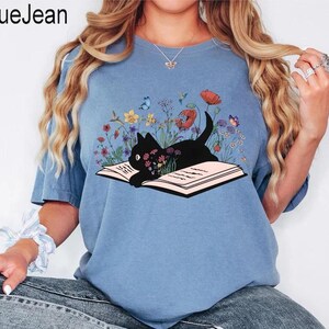 May include: A light blue t-shirt with a black cat reading a book surrounded by flowers. The cat is lying on the open book. The flowers are in shades of pink, purple, yellow, and blue.