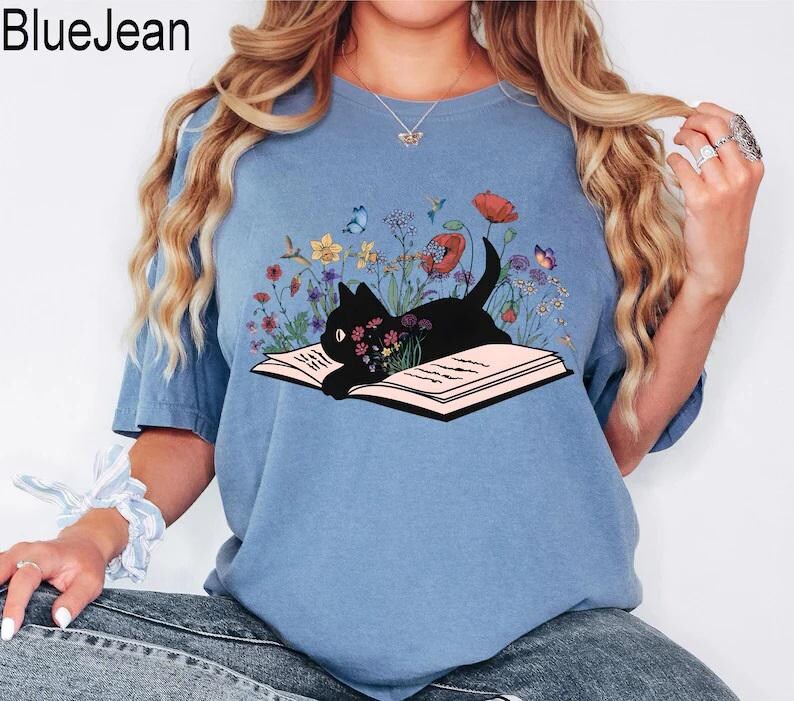 May include: A light blue t-shirt with a black cat reading a book surrounded by flowers. The cat is lying on the open book. The flowers are in shades of pink, purple, yellow, and blue.