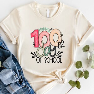 May include: A beige t-shirt with a colorful graphic that says "Happy 100th day of school".