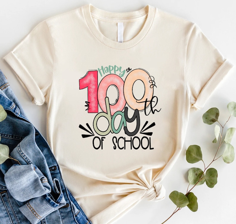 May include: A beige t-shirt with a colorful graphic that says "Happy 100th day of school".