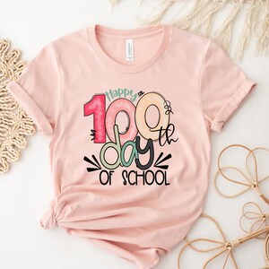 May include: Pink t-shirt with a graphic design that says "Happy 100th day of school". The numbers are colorful and outlined in black.