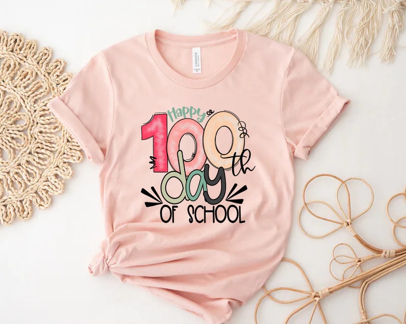 May include: Pink t-shirt with a graphic design that says "Happy 100th day of school". The numbers are colorful and outlined in black.